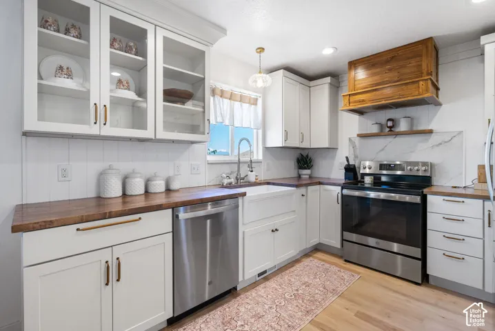 White shaker-style cabinetry with glass-front uppers, wood-finish countertops, stainless steel appliances, a farmhouse sink, and light wood-finish flooring