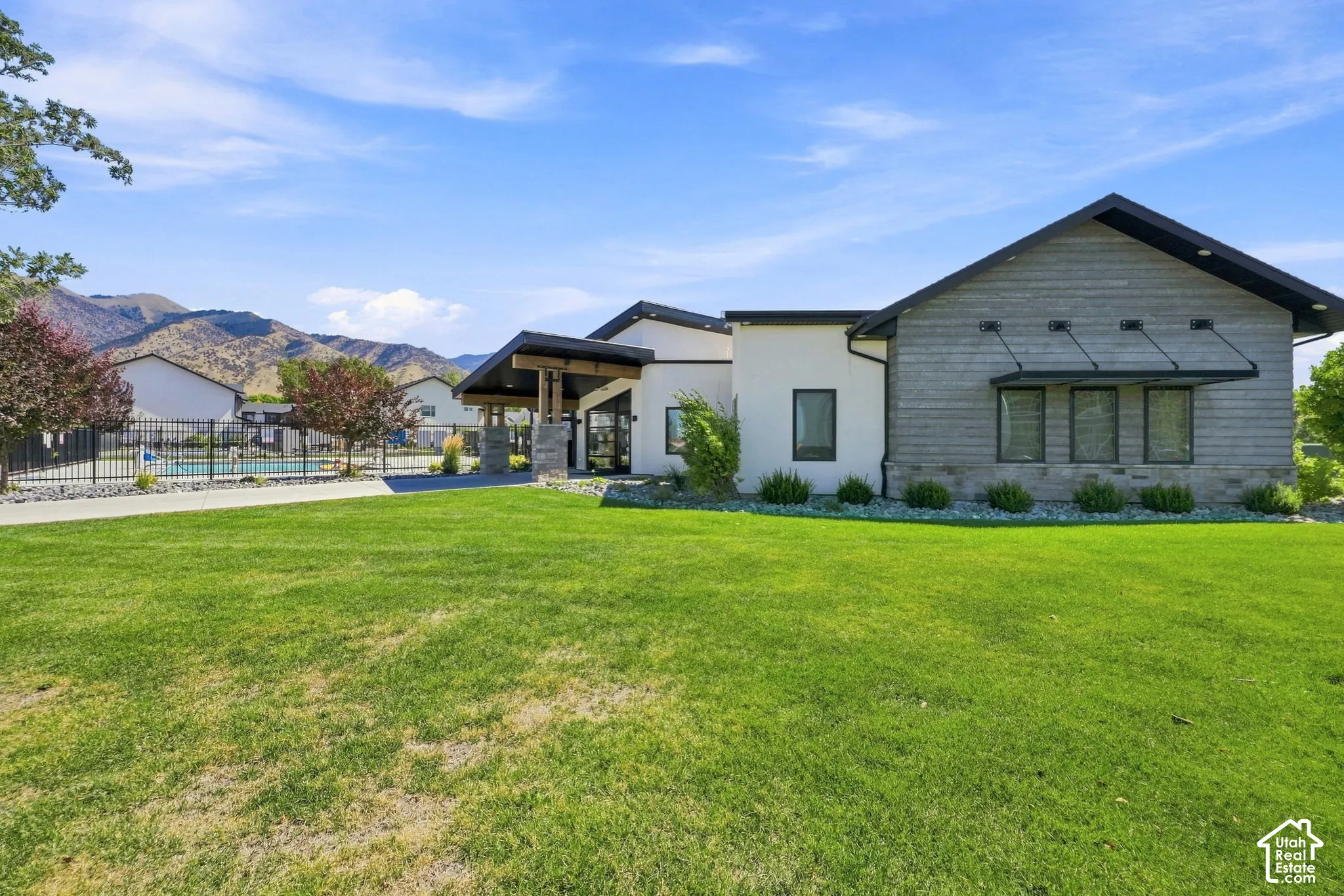 Expansive green space, modern exterior design, covered entryway with wood columns, fenced pool area, and mountain backdrop