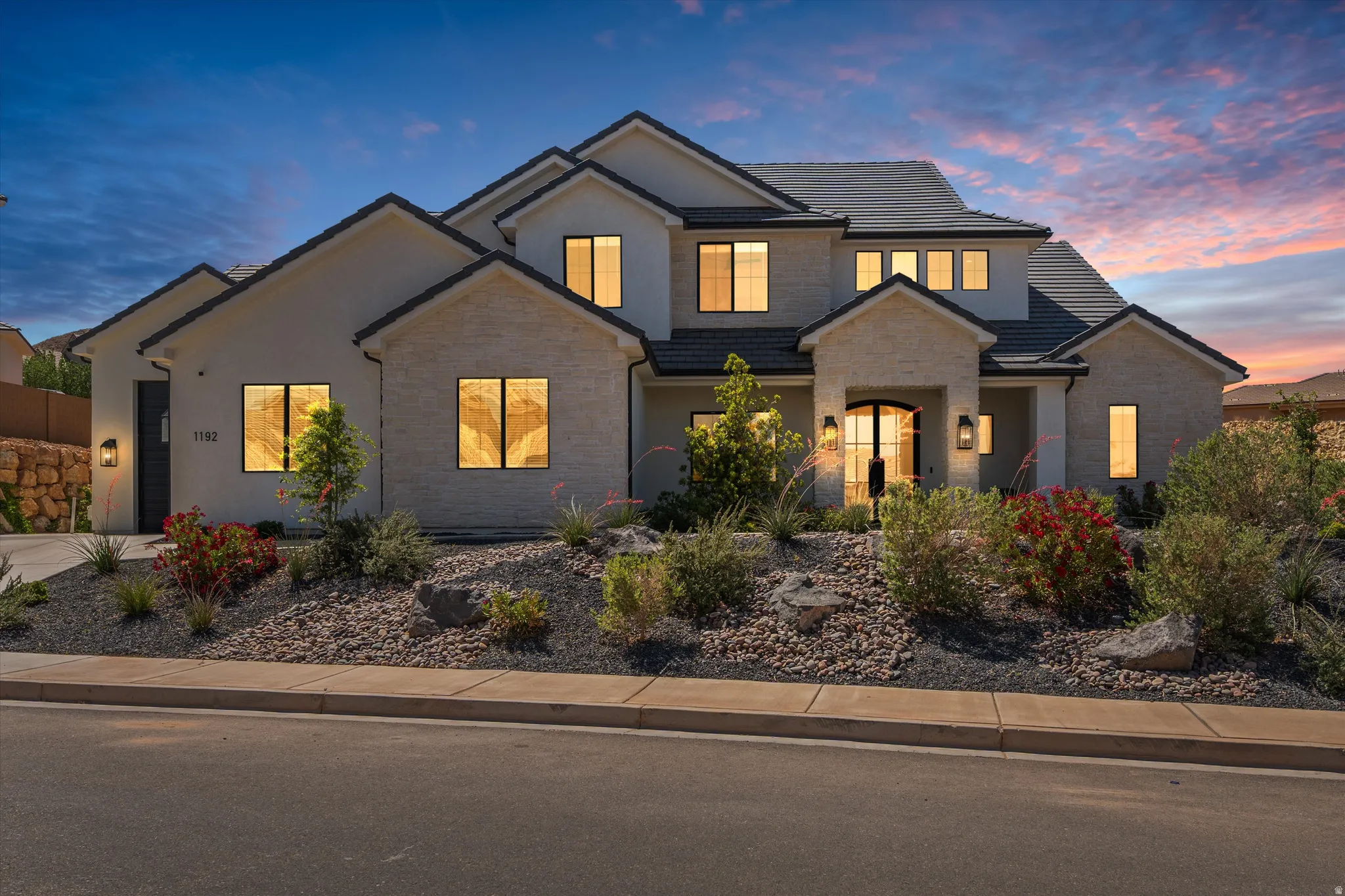 Contemporary exterior featuring light-toned masonry and stucco finishes, a dark tile roof, an arched double front door, and integrated exterior lighting