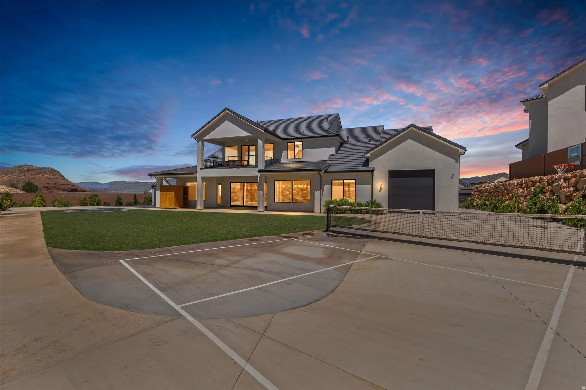 Expansive concrete sport court, attached garage with dark door, covered patio, second-story balcony, and large windows