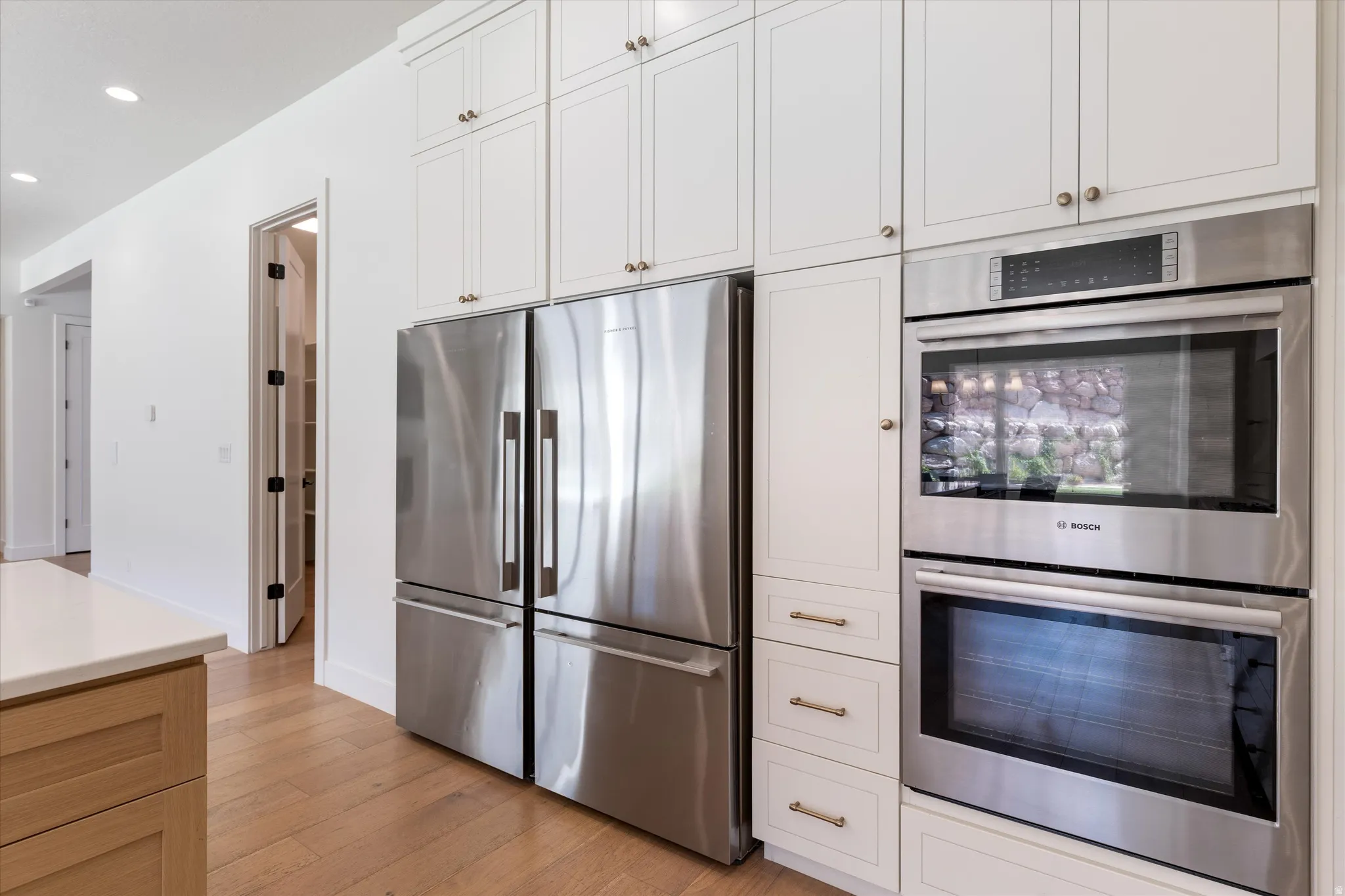 White shaker-style cabinetry with gold-tone hardware, stainless steel built-in double oven, stainless steel refrigerator, light wood-finish flooring, and a pantry with built-in shelving