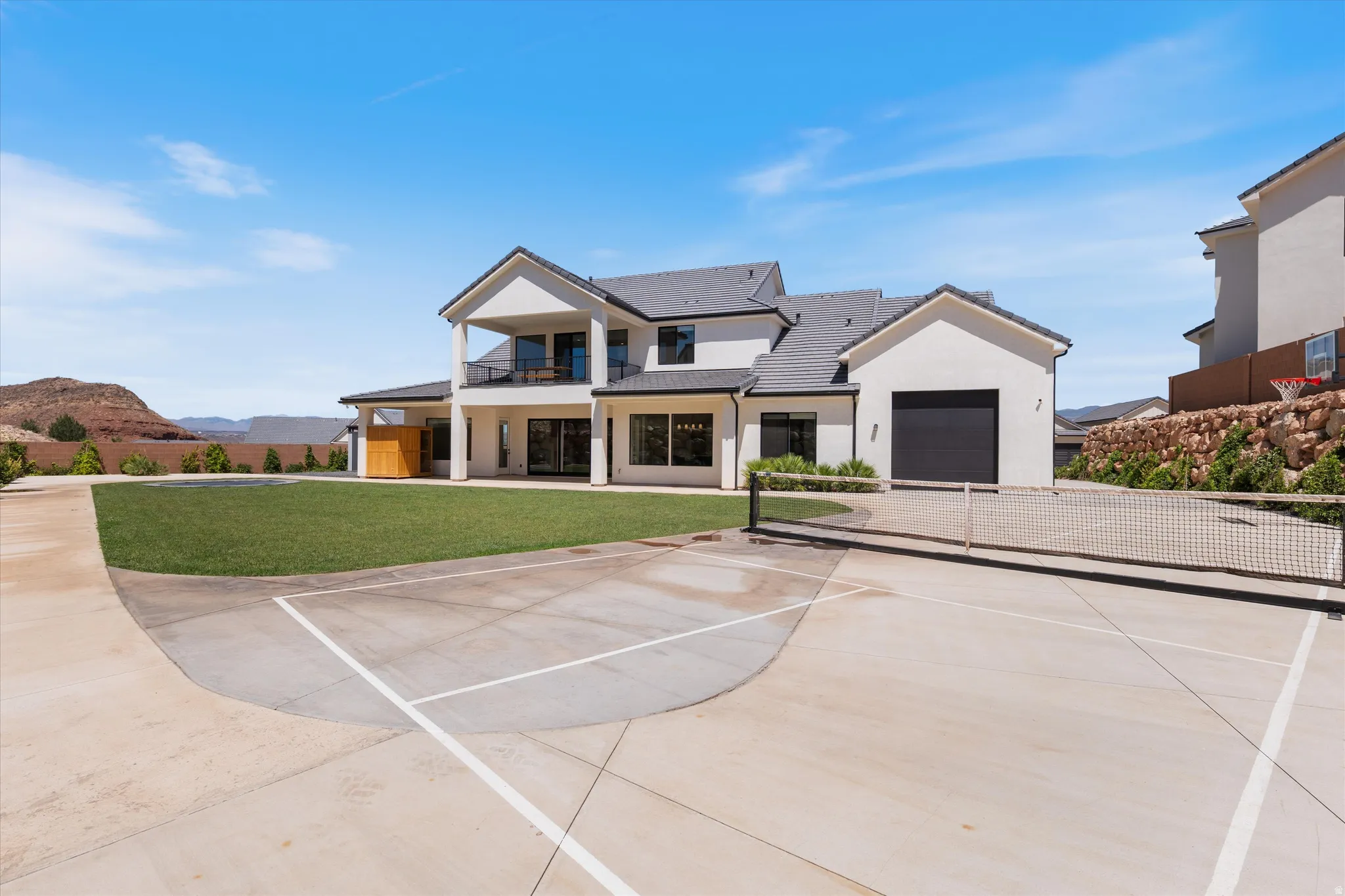 Expansive concrete sport court with a net, broad grass lawn, and a two-story home featuring a balcony, large glass doors, and an attached garage