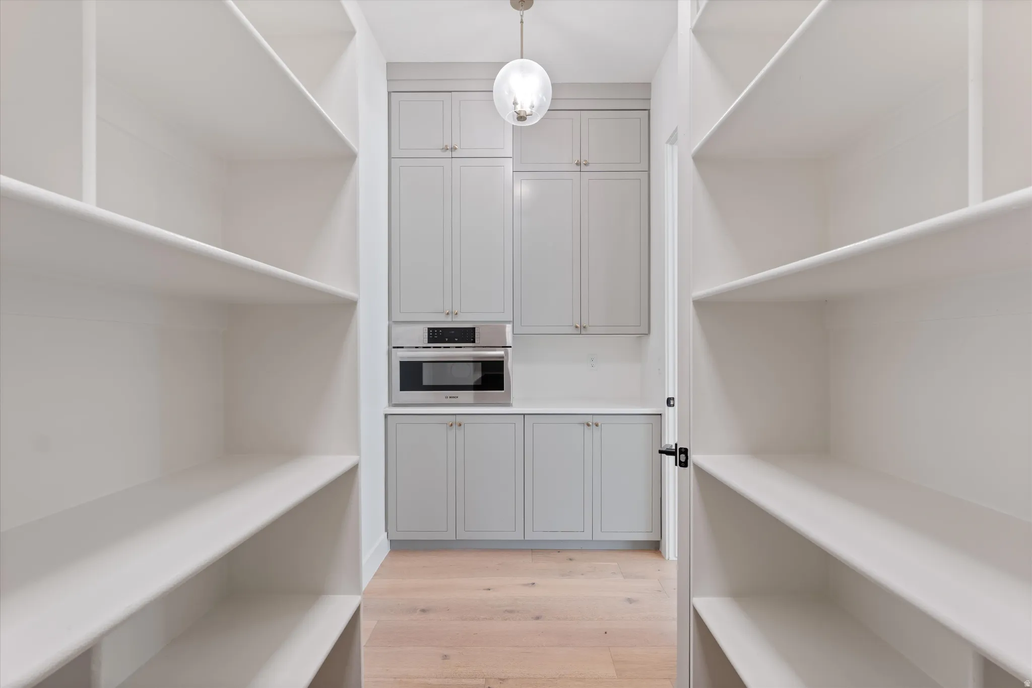 Walk-in pantry featuring extensive built-in shelving, light wood-finish flooring, a built-in stainless steel microwave, light gray cabinetry, and a countertop surface