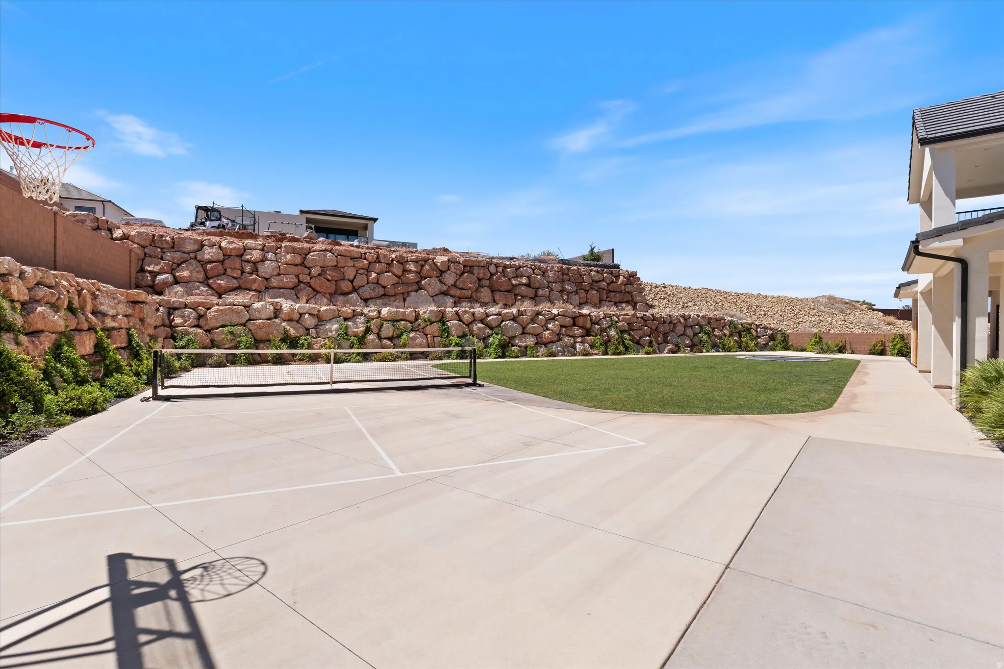 Paved court with game lines and an athletic net, bordered by a manicured lawn and a tiered stone retaining wall
