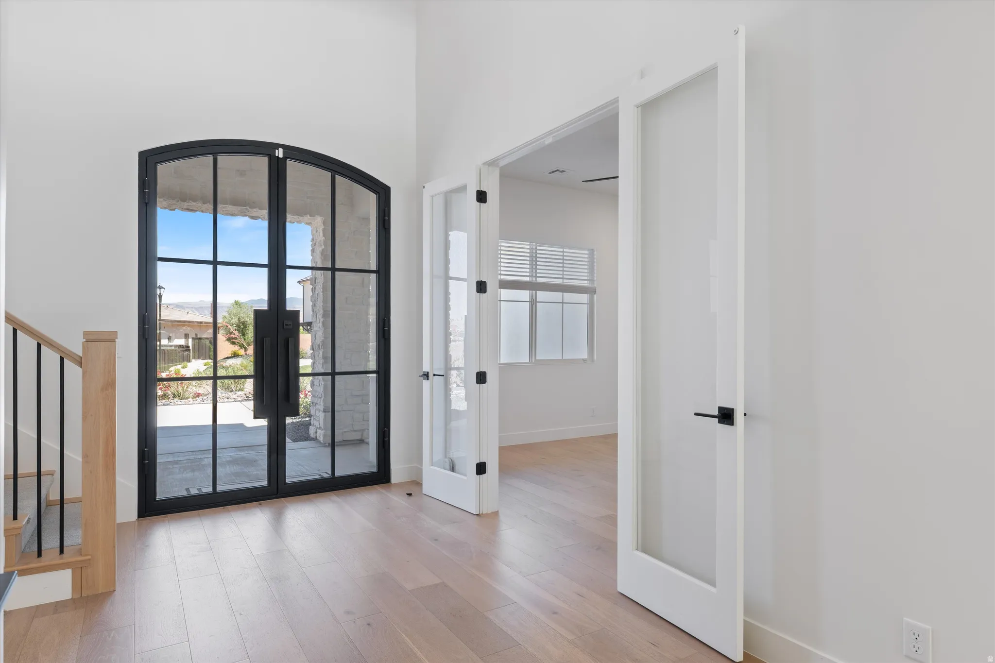 Arched black-frame glass entry doors, wood-finish flooring, white walls, and a wood staircase with black metal balusters