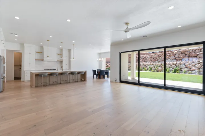 Open concept living area featuring light wood-finish flooring, a large kitchen island with pendant lighting, white cabinetry, and expansive multi-panel sliding glass doors leading to a private outdoor space