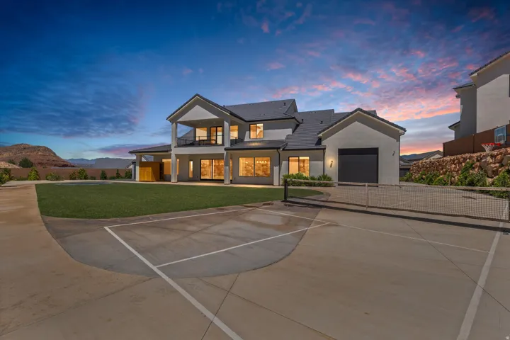 Expansive concrete sport court, attached garage with dark door, covered patio, second-story balcony, and large windows