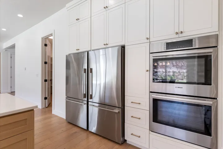 White shaker-style cabinetry with gold-tone hardware, stainless steel built-in double oven, stainless steel refrigerator, light wood-finish flooring, and a pantry with built-in shelving
