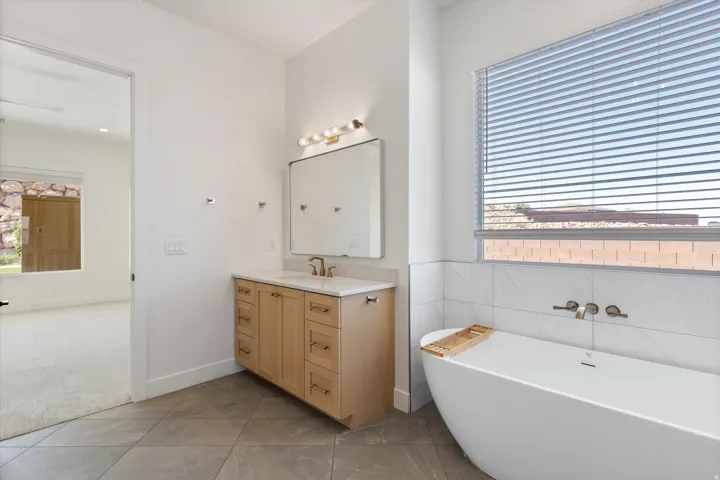 Freestanding soaking tub with wall-mounted bronze faucet, wood-finish vanity with countertop sink, large mirror, and overhead vanity lighting