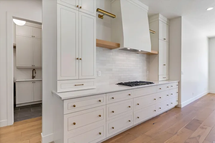 Integrated gas cooktop, white subway tile backsplash, custom range hood, extensive white cabinetry with brass hardware, and wood-finish flooring