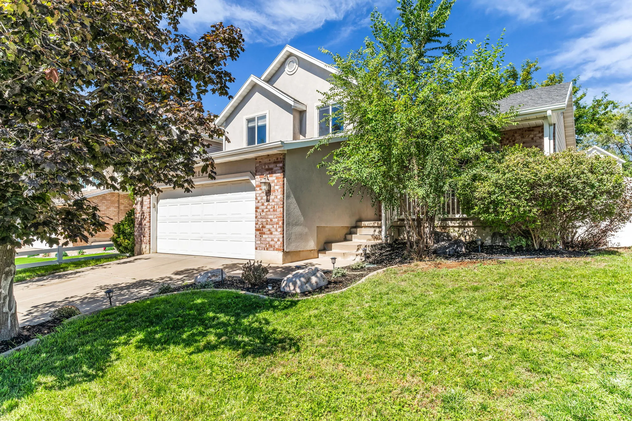 Exterior featuring stucco and brick accents, an attached garage, a concrete driveway, a front lawn, and mature trees