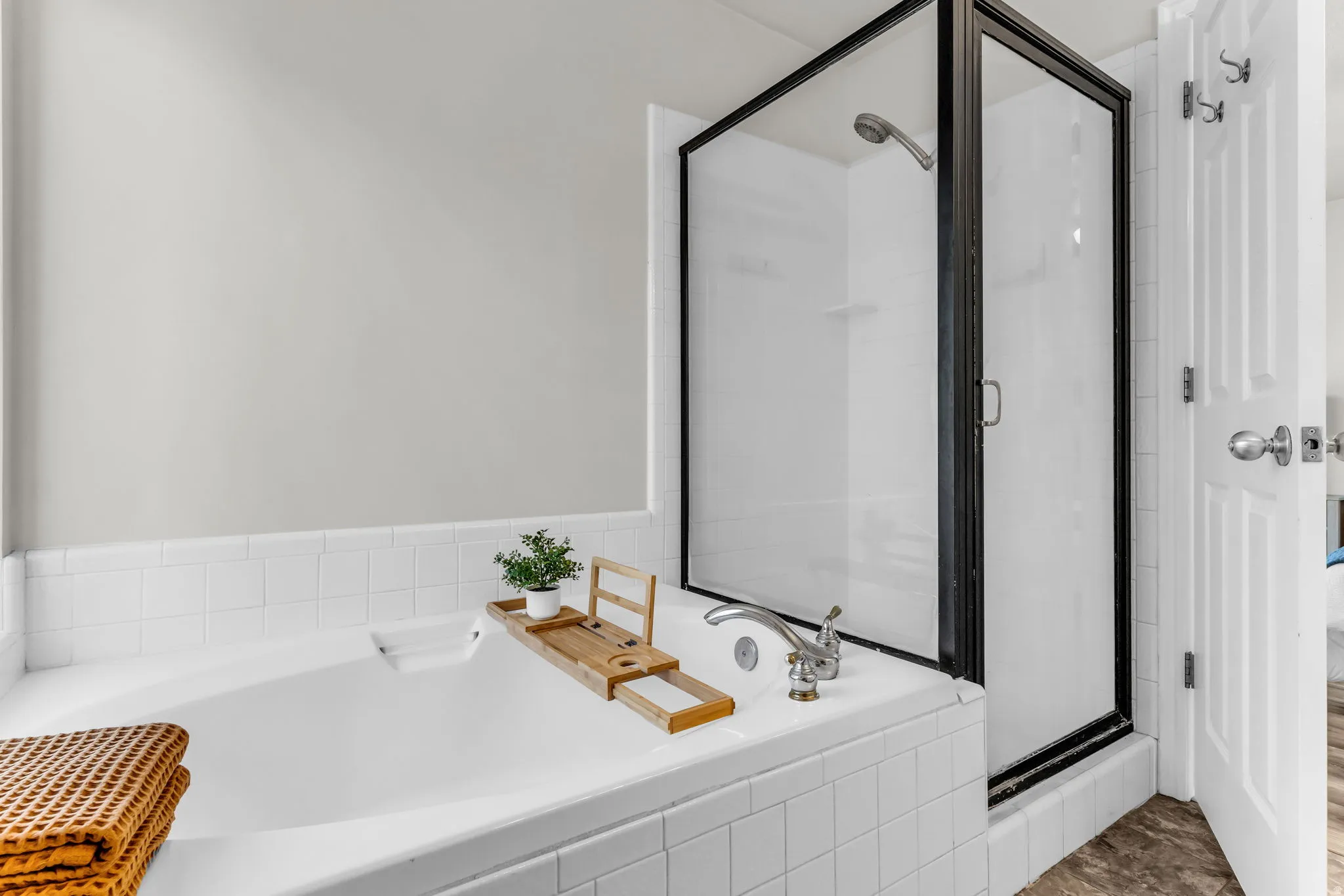 Built-in soaking tub with white tile surround, adjacent to a glass-enclosed shower with a black frame