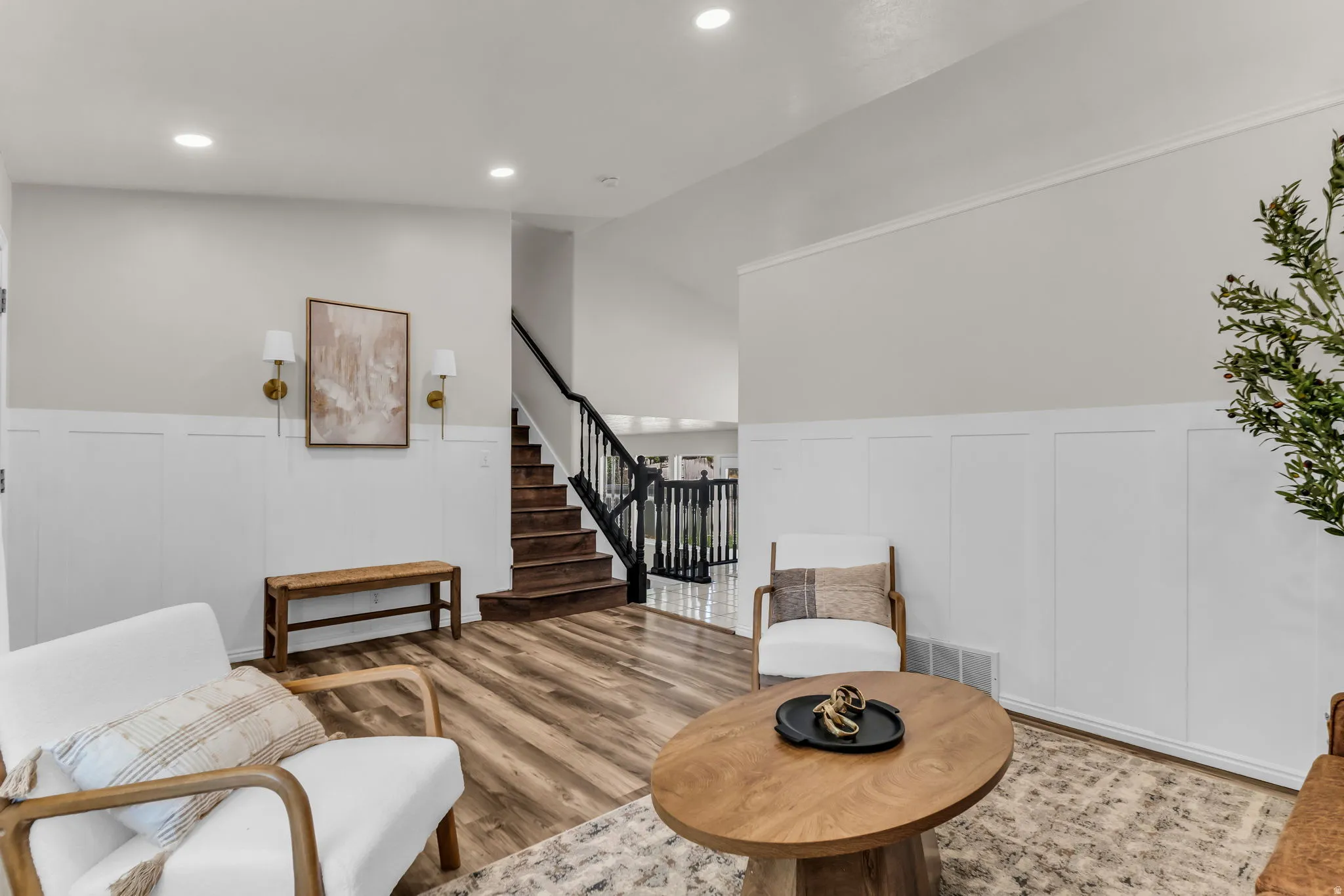 Recessed lighting illuminating wood-finish flooring, white wainscoting, and a staircase with dark wood treads and balusters