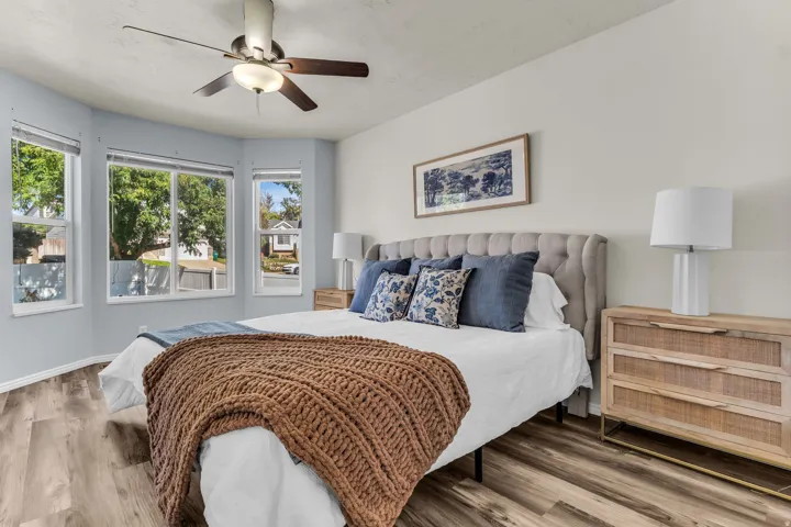Bay window with three large windows, a ceiling fan with integrated lighting, wood-finish flooring, and a tufted upholstered headboard