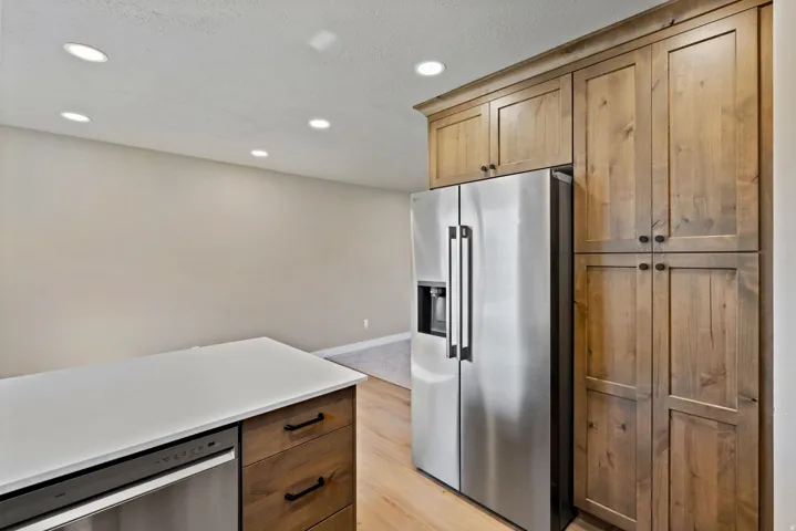 Stainless steel refrigerator, integrated dishwasher, light wood-finish cabinetry, white countertop, recessed lighting