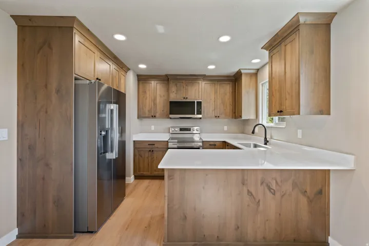 Modern kitchen featuring warm wood cabinetry throughout, stainless steel appliances including a French-door refrigerator and electric range, white quartz countertops with waterfall island edge, recessed ceiling lighting, wood-finish flooring, and a underm