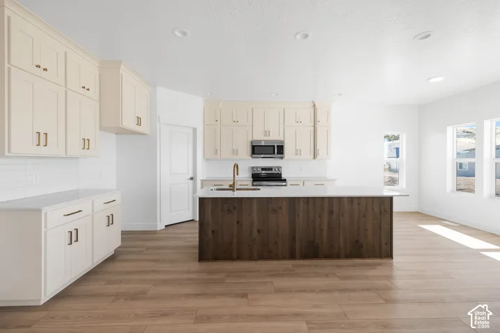 Kitchen featuring light-toned cabinetry, a spacious island with wood-finish paneling, stainless steel appliances, a tile backsplash, and light wood-finish flooring