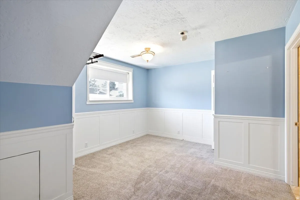 Recessed window with roller shade, wainscoting, textured carpet, ceiling fan with light fixture, and sloped ceiling
