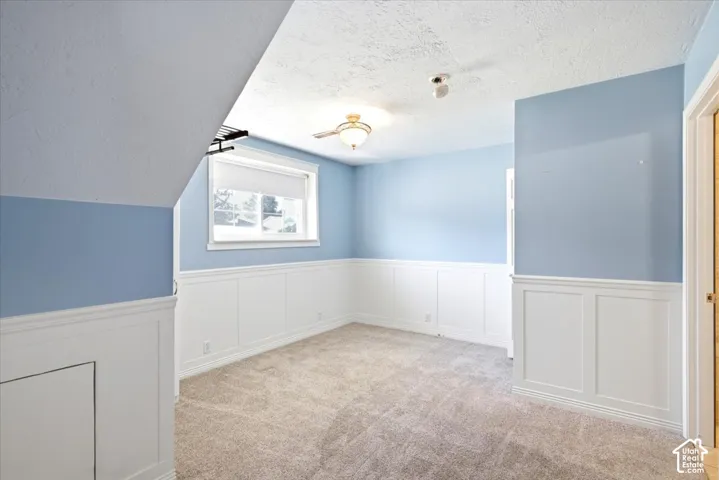 Recessed window with roller shade, wainscoting, textured carpet, ceiling fan with light fixture, and sloped ceiling