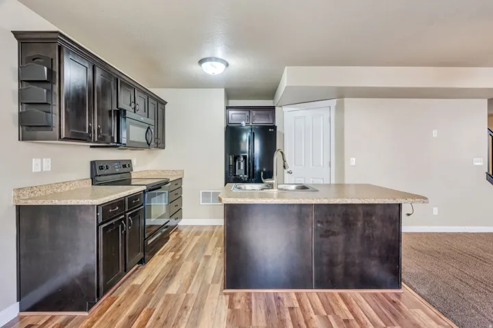 Kitchen featuring dark wood-finish cabinetry, light-toned countertops, a center island with a double basin sink, stainless steel appliances, and wood-finish flooring