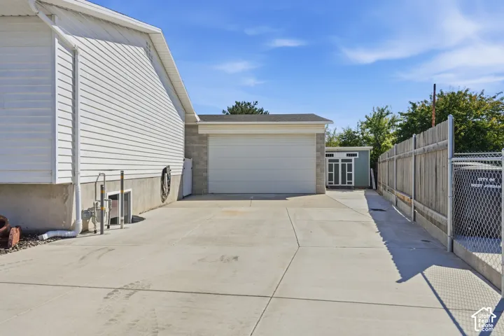 Expansive concrete driveway, white horizontal siding, garage with automatic door, wood privacy fence, and chain-link fencing