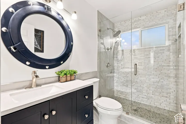 Dark-toned vanity with a white countertop and integrated sink, round mirror with accent lighting, glass-enclosed shower with a rainfall showerhead, and subway tile shower surround