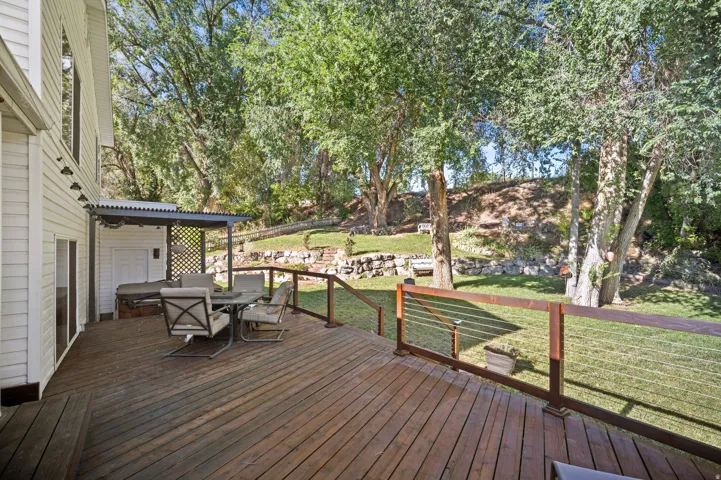 Spacious wood deck with cable railing, featuring a covered area and direct access to a tiered backyard with stone retaining walls and mature trees