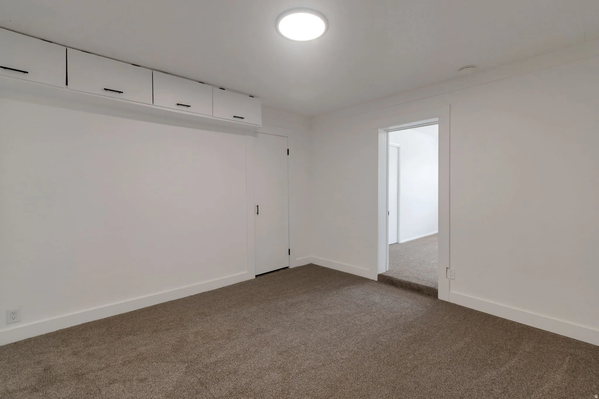 White walls with built-in overhead cabinetry featuring dark hardware, a flush-mount ceiling light, and brown carpeting