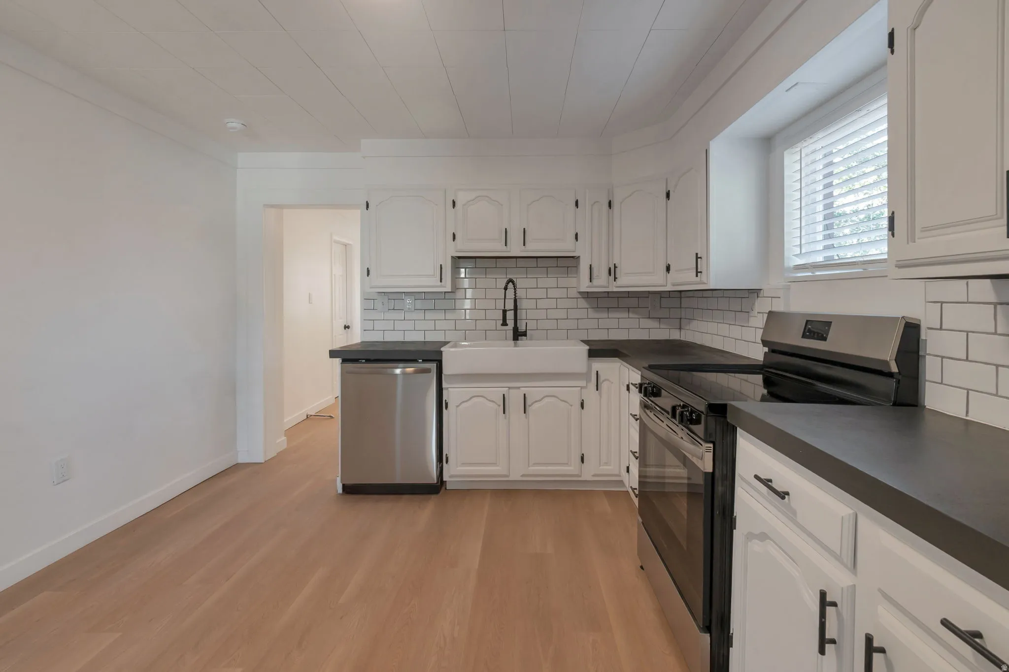 Kitchen featuring light wood-finish flooring, white cabinetry with dark hardware, dark countertops, white subway tile backsplash, and a farmhouse-style sink