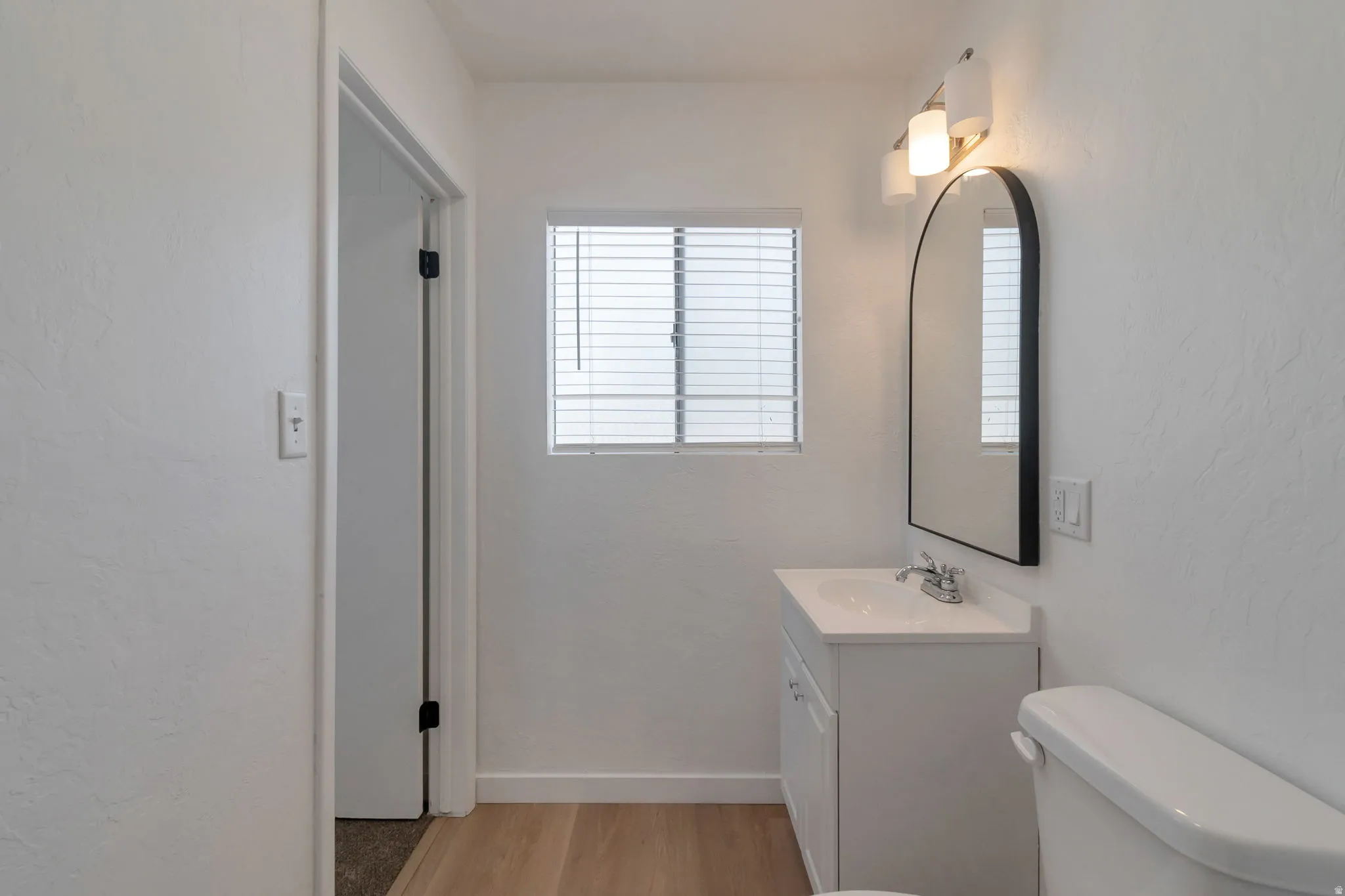 White vanity with an integrated sink, arched mirror, three-light vanity fixture, and wood-finish flooring