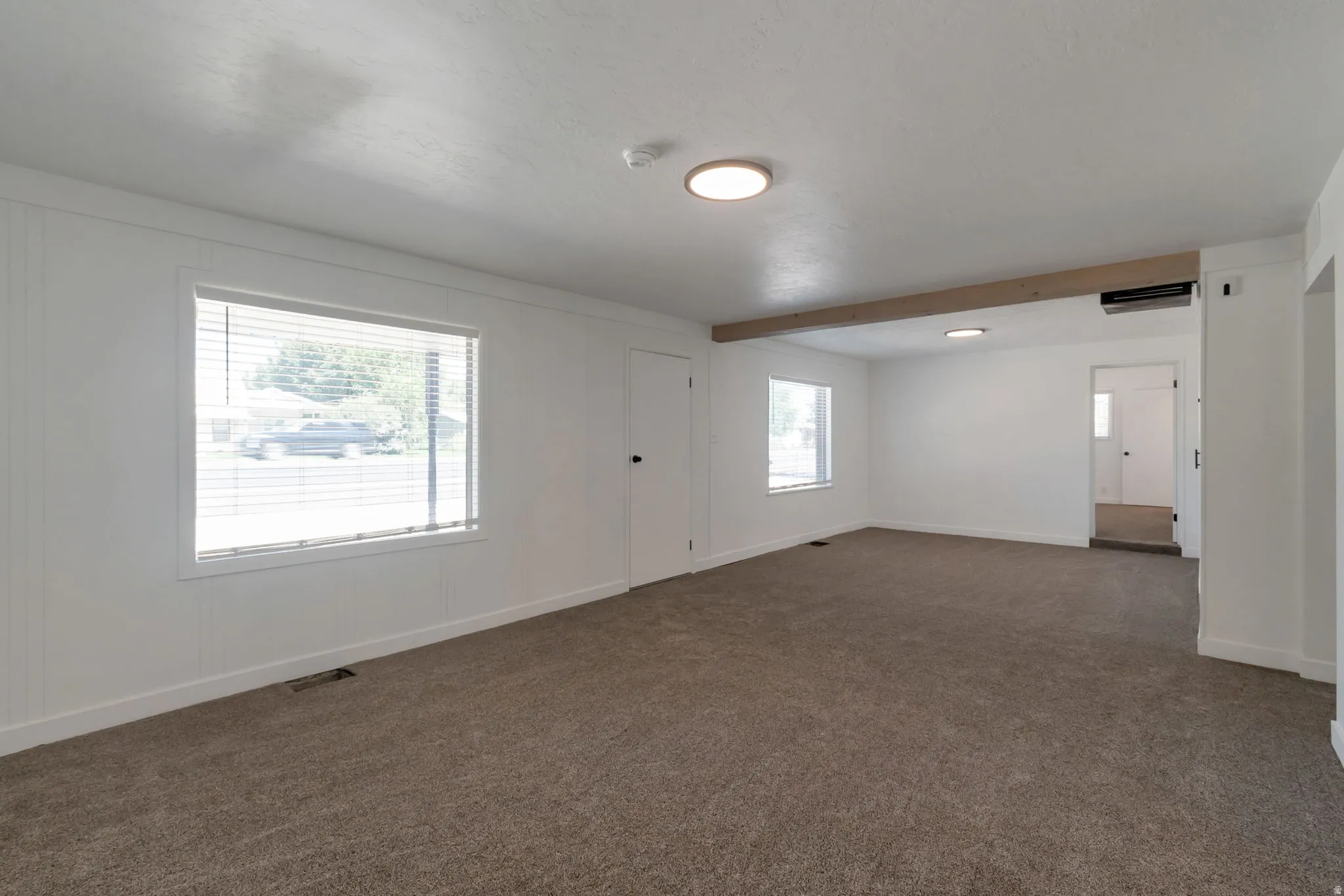 Carpet flooring, multiple windows with blinds, flush-mount lighting, and an exposed wood beam