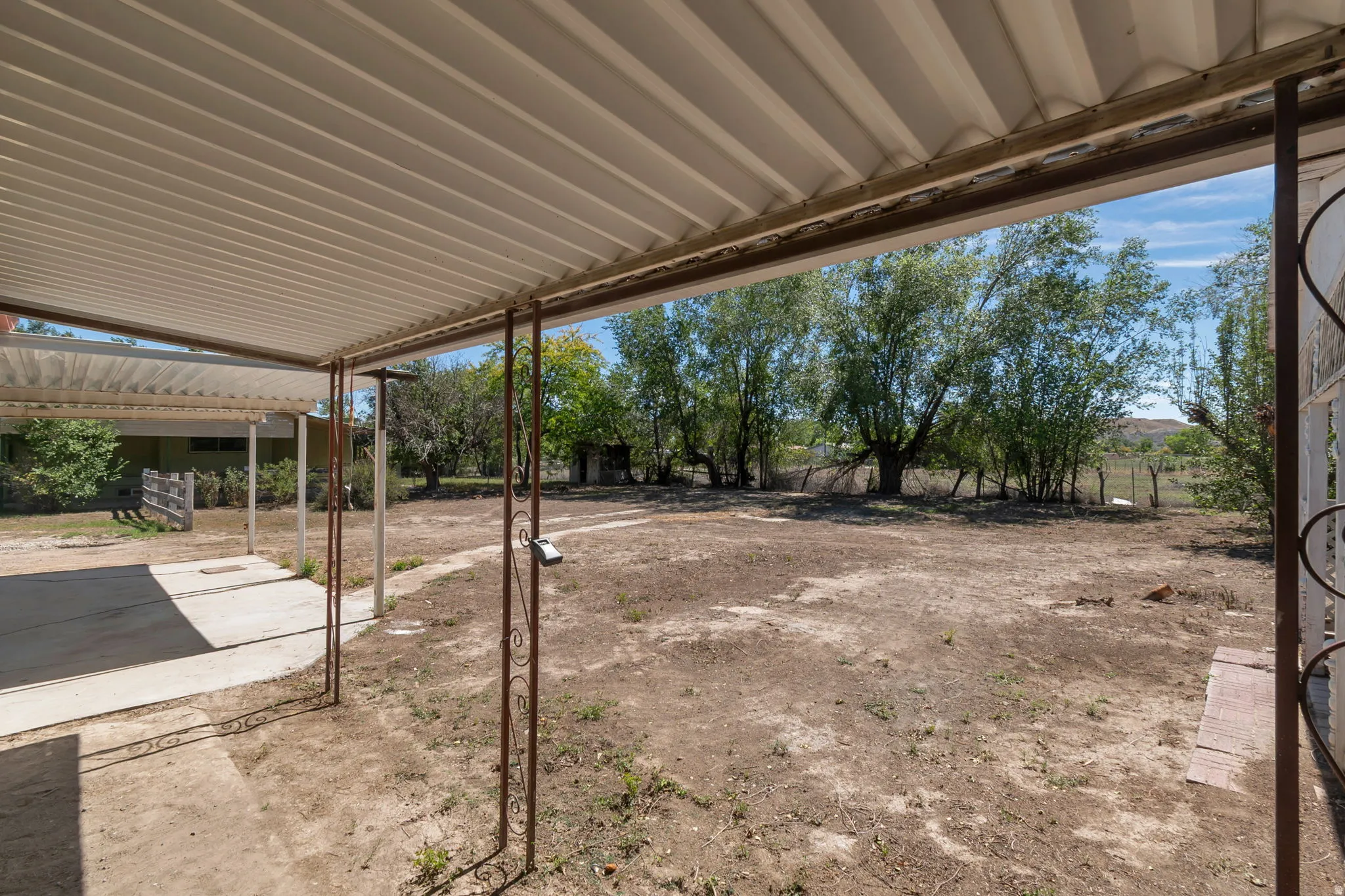 Expansive outdoor area featuring a covered patio with corrugated metal roof, concrete slab, and wrought iron supports