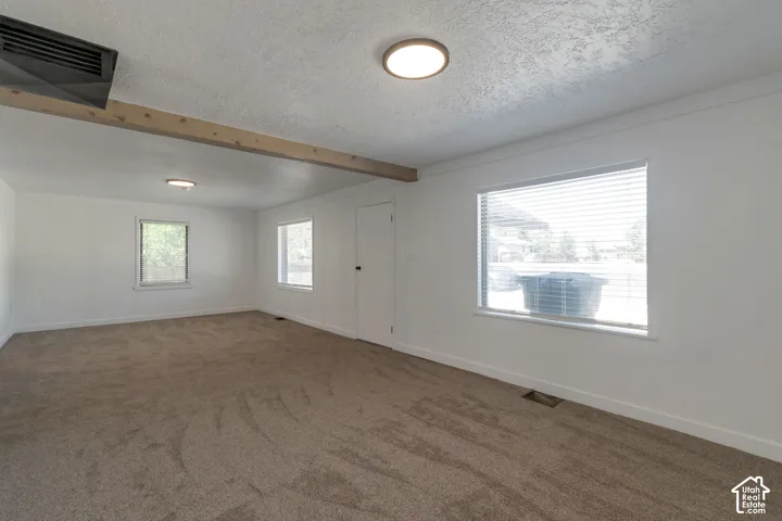 Spacious room featuring new carpeting, white walls, multiple windows with blinds, and exposed ceiling beams