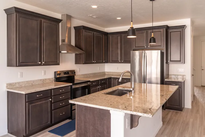 Kitchen featuring dark wood-finish cabinetry, light-toned countertops, stainless steel appliances, a center island with an undermount sink, and pendant lighting