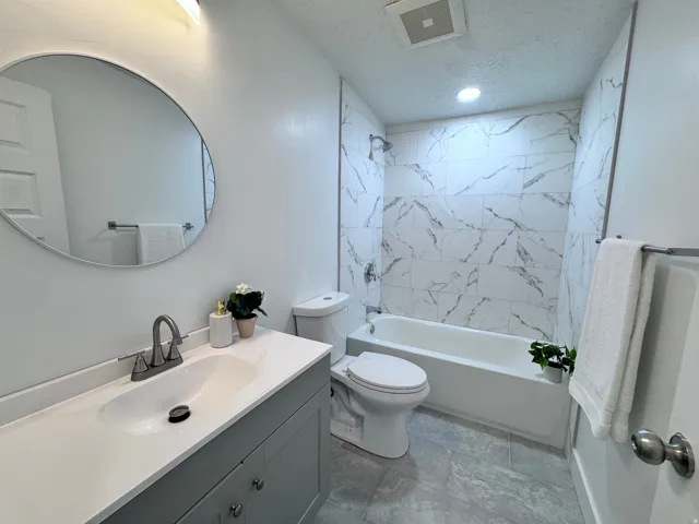 Vanity with a white countertop and undermount sink, circular mirror, and patterned wall tile in the bath/shower enclosure