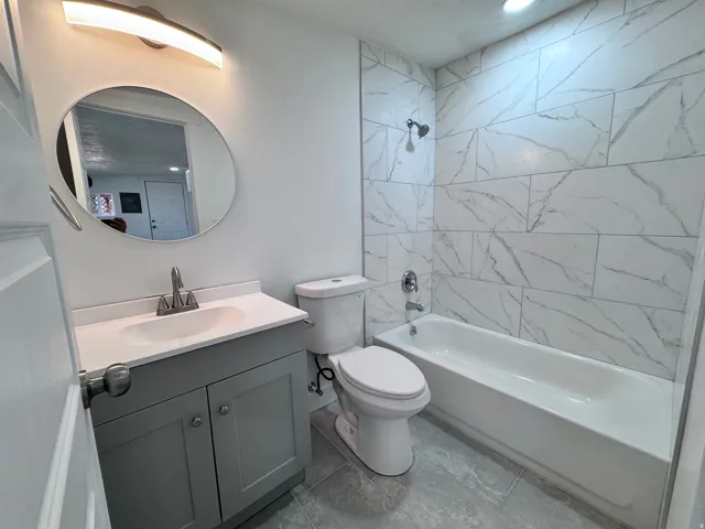 White subway tile shower surround, gray wood-finish vanity with white countertop, round mirror, and tile flooring