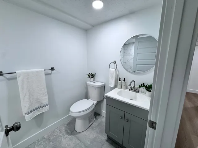 Bathroom featuring gray tile flooring, a white toilet, a vanity with a white countertop and gray cabinetry, and a circular wall mirror