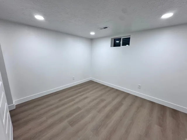 Recessed ceiling lighting illuminates a room featuring wood-finish flooring, white walls, and a window with blinds