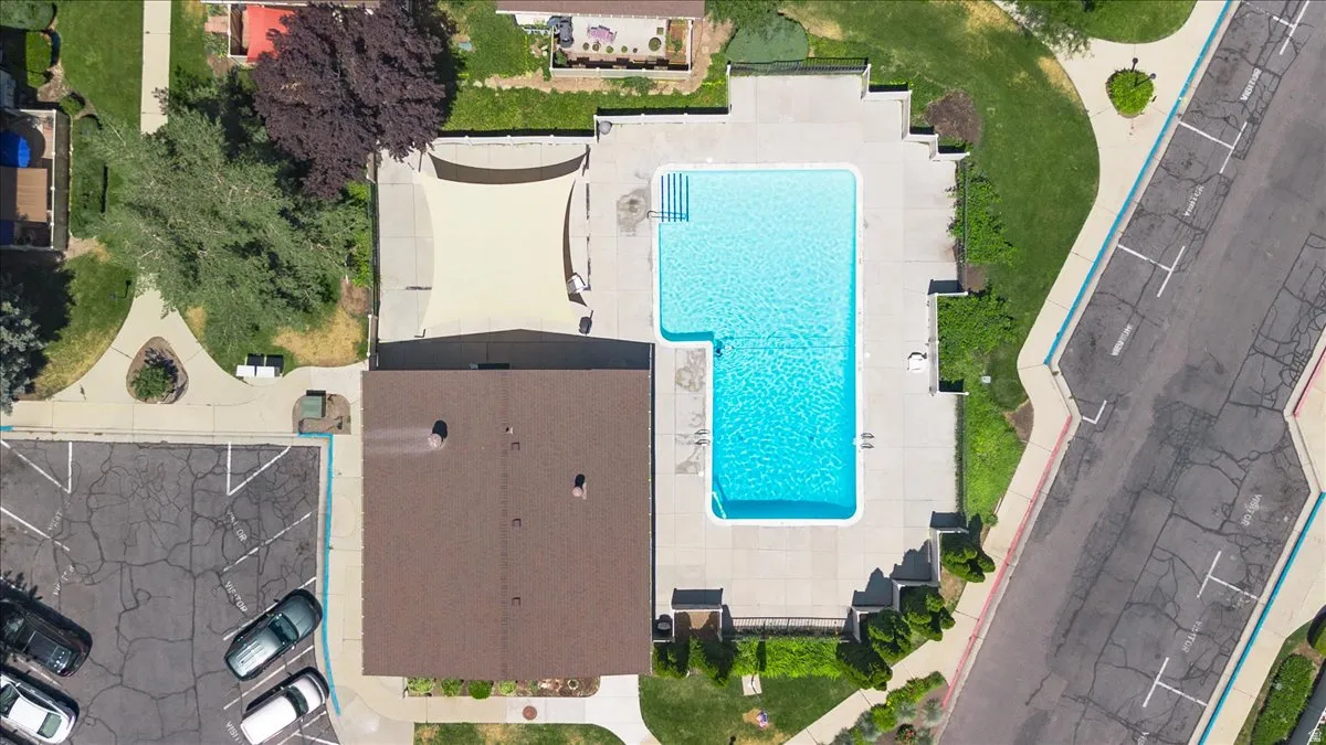 Community pool with a surrounding concrete deck, adjacent building with a brown roof, shaded patio area, and nearby parking