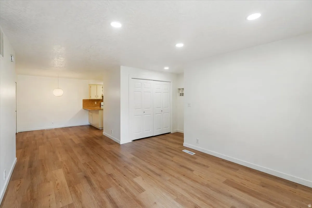 Recessed lighting, wood-finish flooring, white walls, built-in closet with bifold doors, and a partial view of a kitchen with light cabinetry