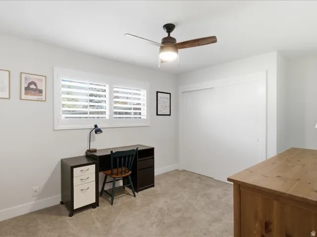 Neutral wall paint, plush carpet flooring, ceiling fan with integrated lighting, window with white plantation shutters, and a closet with sliding doors