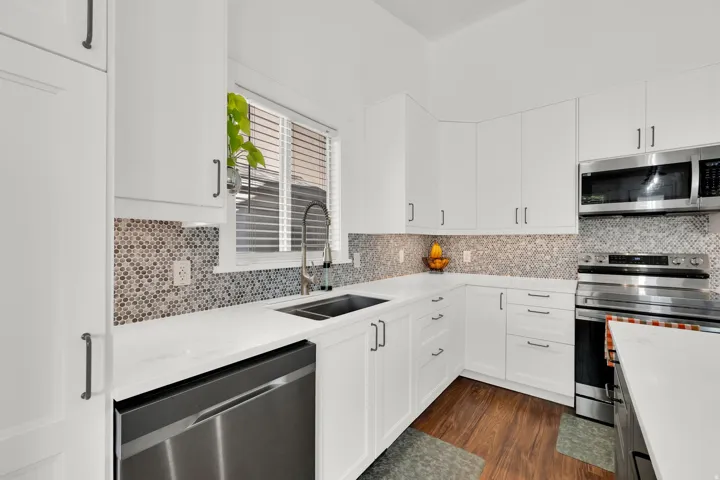 White cabinetry with dark hardware, stainless steel appliances, patterned tile backsplash, wood-finish flooring, and a double basin sink