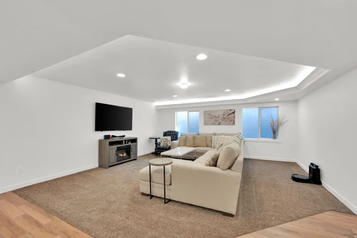 Sunken tray ceiling with perimeter lighting, recessed ceiling lights, wall-mounted television, integrated electric fireplace, and multiple windows