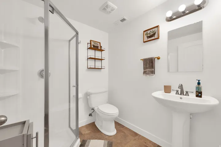Glass-enclosed shower with built-in shelving, pedestal sink, wall-mounted mirror, and overhead vanity lighting