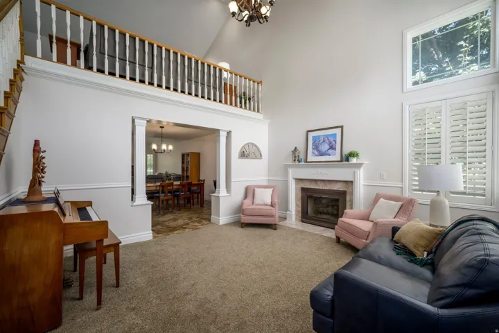 Vaulted ceiling with an upper window, prominent fireplace with a light-toned surround, plantation shutters, open archway with decorative columns, and a staircase with wood railings