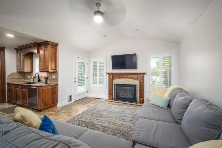 Vaulted ceiling with a ceiling fan, gas fireplace with a wood-finish mantel and stone surround, wood-finish cabinetry, built-in dishwasher, and a glass-paneled door