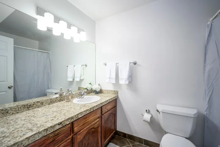 Full-width vanity mirror, six-light fixture, wood-finish cabinetry, speckled countertop, and a white toilet