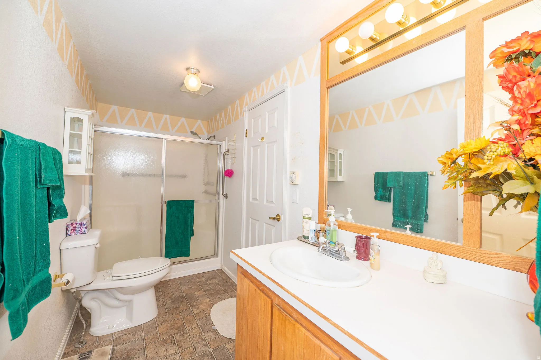 Vanity with integrated sink and wood-finish cabinetry, framed mirror with overhead lighting, shower enclosure with sliding glass door, white toilet, and tile flooring