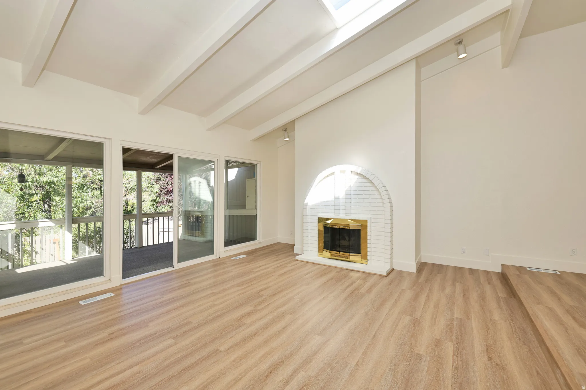 Vaulted ceiling with exposed beams, wood-finish flooring, white brick fireplace with brass surround, multiple glass sliding doors, and a skylight