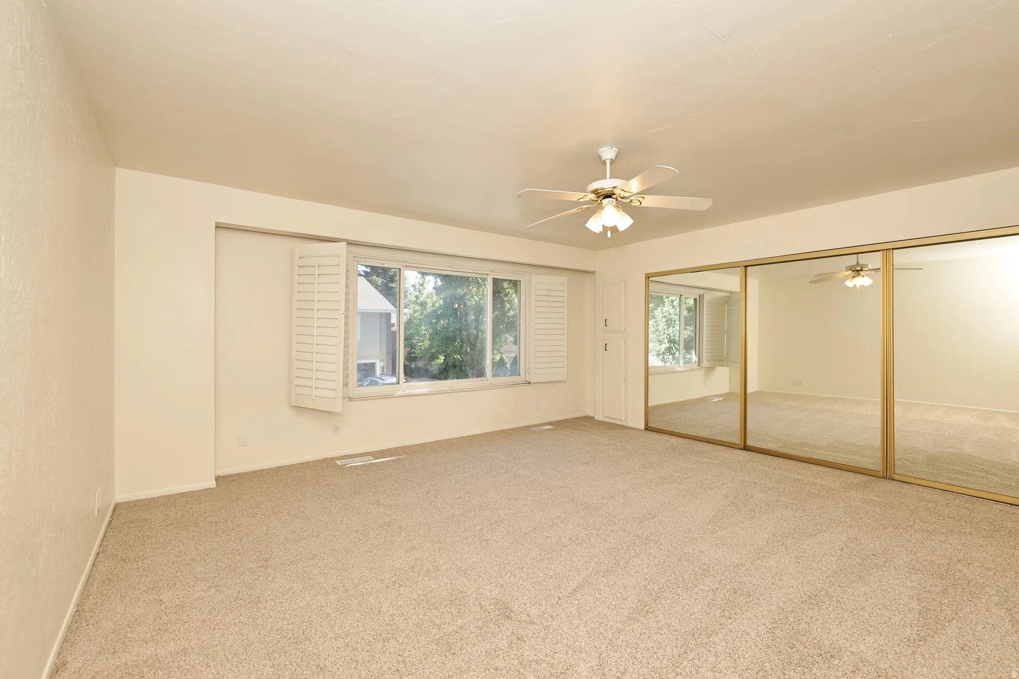 Carpeted room featuring a ceiling fan with light fixture, large windows with white shutters, and mirrored sliding closet doors